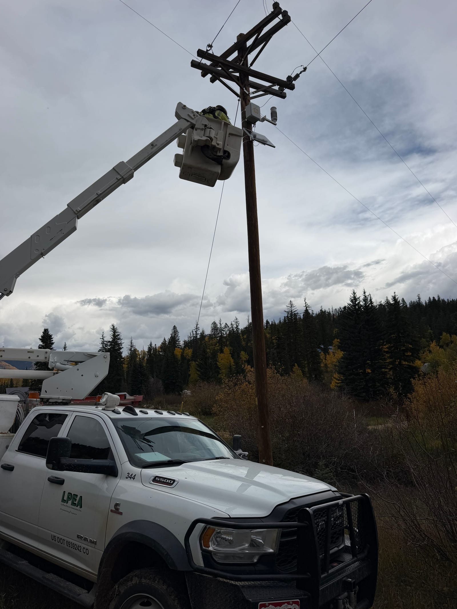 Lineman in an LPEA bucket truck installing a WatchPost weather station near the top of a utility pole, evergreen forest behind