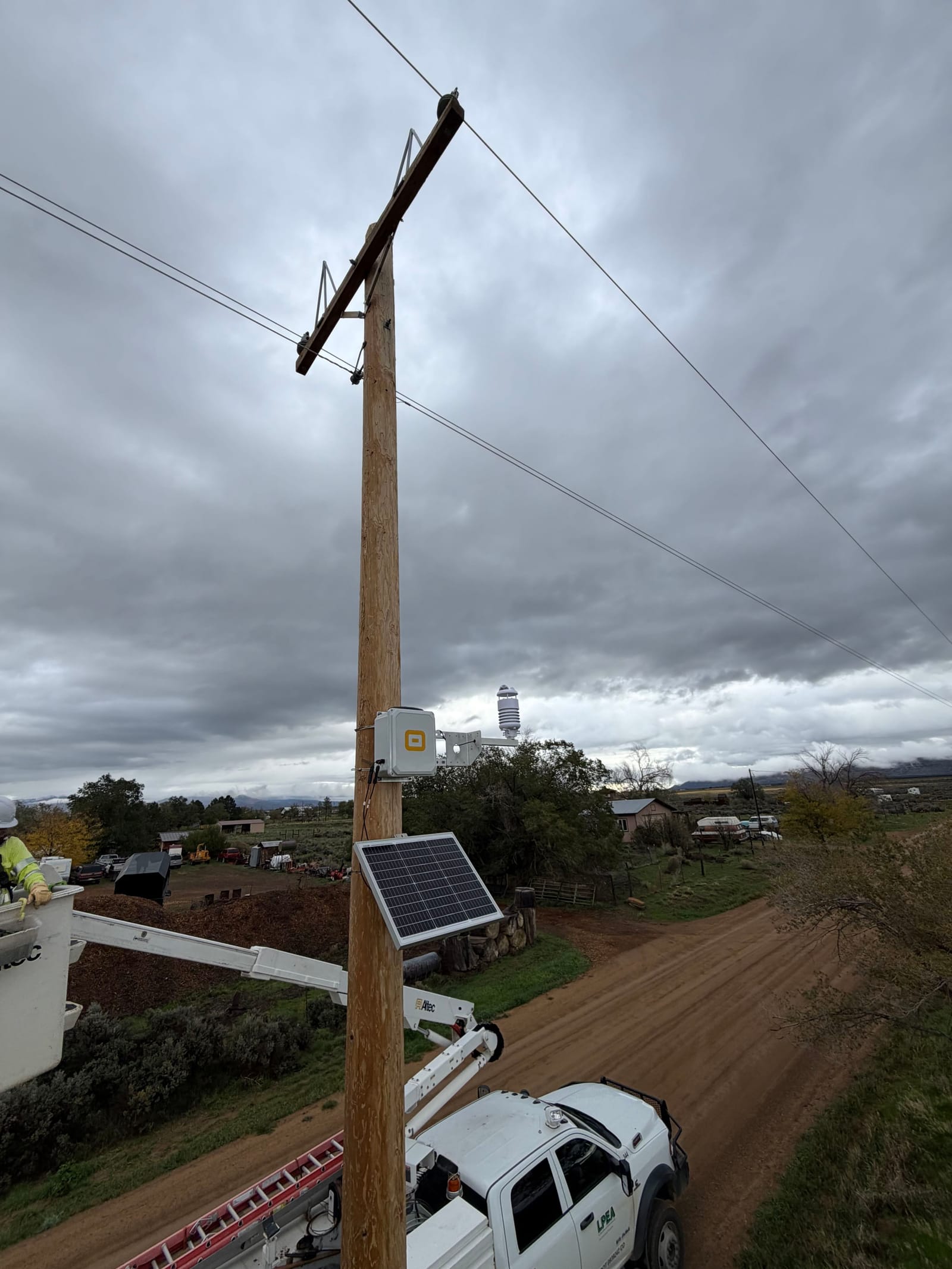 LPEA bucket crew beside a utility pole fitted with a solar-powered WatchPost station above a dirt ranch road under storm clouds